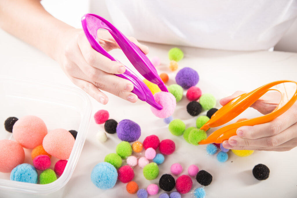 A children holding soft pom pom balls with large plastic tweezers as part of a craft and handeye coordination activity as directed by their childrens occupational therapist